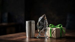 A personalized tumbler, glass award, and green-ribbon gift box on a dark desk.